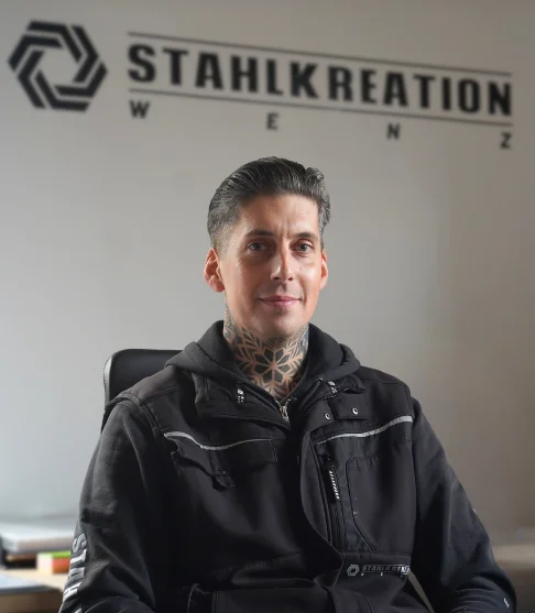Daniel Wenz in Arbeitsjacke vor Stahlkreation-Logo im Büro